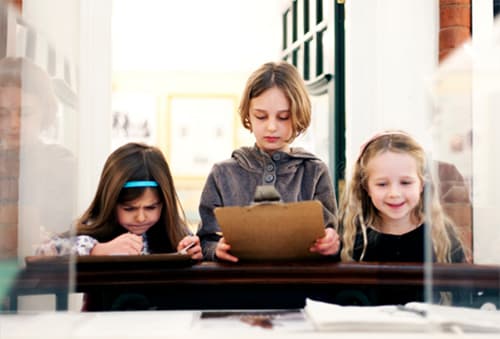 Three young children looking at an exhibition with a clipboard in hand.
