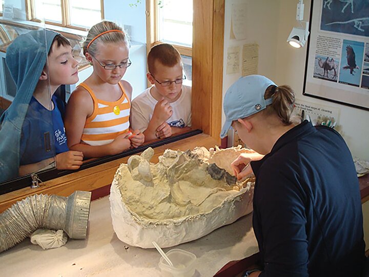 Three children watch a researcher excavate a fossil inside a field jacket in the working visible lab of Ashfall Fossil Beds.
