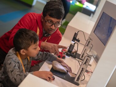 A father and son examine a large rock under a microscope displaying to a large monitor